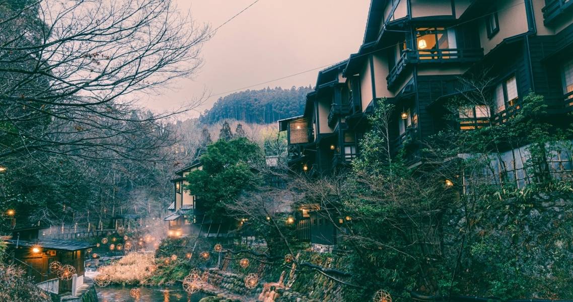 Kurokawa Onsen im Winter: Das warme Licht der Bambuslaternen spiegelt sich auf dem Wasser, am Horizont sind Berge zu sehen